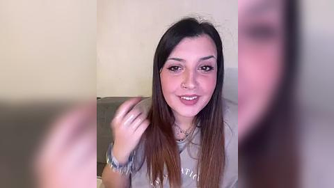 Media: Video of a young woman with long, straight, dark brown hair, light skin, and brown eyes, smiling, wearing a gray t-shirt, in a plain beige room.