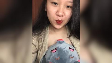 Media: Video of an Asian woman with long black hair, light skin, and red lipstick puckering her lips. She wears a beige cardigan over a plush toy resembling a sleeping Pikachu, with closed eyes and a small red nose. Background is blurred.