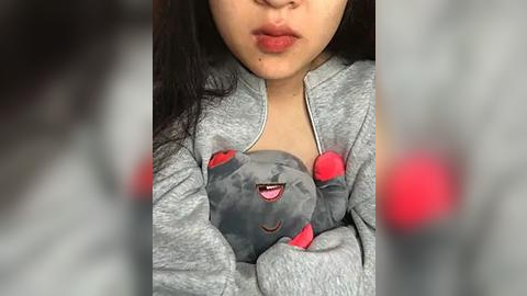 Media: Video of a young Asian woman with long black hair, wearing a gray zip-up hoodie with a plush gray teddy bear with red ears, smiling.