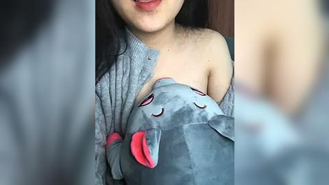 Media: A video of a young woman with fair skin and long black hair, wearing a grey off-shoulder cardigan, cuddling a plush grey elephant with pink ears. The background is blurry, featuring a wooden door.