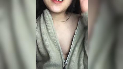 Media: Video of a young Asian woman with fair skin, wearing a light gray zip-up hoodie, partially unzipped, revealing a hint of her breast. Her dark hair falls over her shoulders.