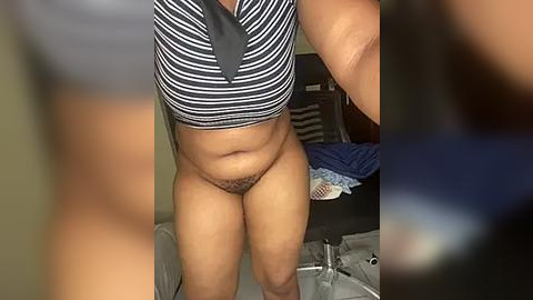 Media: Video of a woman in a black and white striped crop top, exposing her stomach and pubic hair, standing in a bedroom with unmade bed and clothes on the floor.