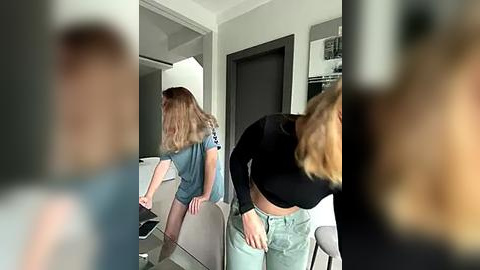 Media: Video of a woman with long blonde hair in a black crop top and light jeans adjusting her jeans in a modern, sparsely furnished living room with light grey walls and a black door.