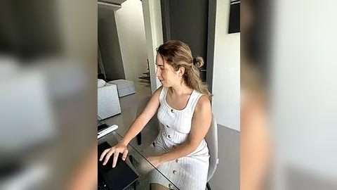 Media: Video of a young woman with light skin and brown hair, wearing a white sleeveless dress, sitting at a modern, sleek black table in a minimalist, well-lit room with white walls and a black door.