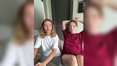 Media: Video of two women with fair skin, shoulder-length blonde hair, in casual white and red shirts, sitting on a sofa in a modern living room. Background includes a white door and black wall.