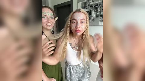 Media: Video of two young women with elf ears, one with long blonde dreadlocks, the other with brown hair, smiling, indoors near a black-and-white cityscape print.