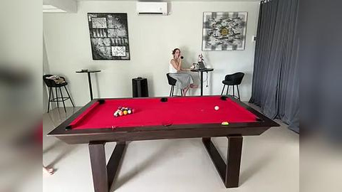 Media: Video of a modern, minimalist room with a pool table, red felt, and black legs. A woman in a sleeveless top sits at a small table, and two black stools are nearby.