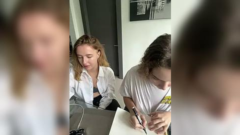 Media: Video of two young women in casual attire, one with blonde hair, the other with curly brown hair, seated at a desk, writing and reading. Background features a black and white framed picture.