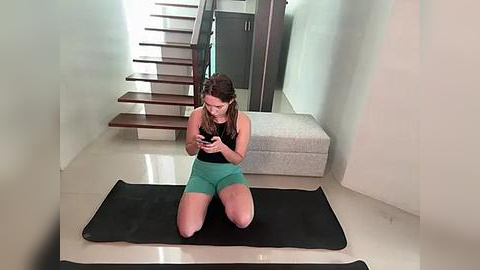 Media: Video of a woman with light skin, brown hair, in a black tank top and green shorts, kneeling on a yoga mat, focused on her smartphone in a modern, minimalist room with a wooden staircase and light-colored walls.