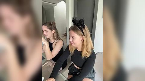 Media: Video of two young women in casual attire, one with a bun and a black top, the other with blonde hair, wearing a black top and shorts, sitting on a glass table, in a modern, minimalist room.