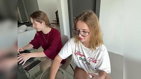 Media: Video of two young women with glasses, one in a maroon top, the other in a white Coca-Cola shirt, playing a video game at a glass table in a modern living room.