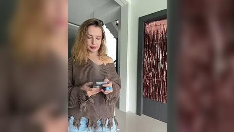 Media: Video of a blonde woman in a distressed brown sweater, light blue jeans, holding a phone, standing in a modern room with a tasseled curtain.