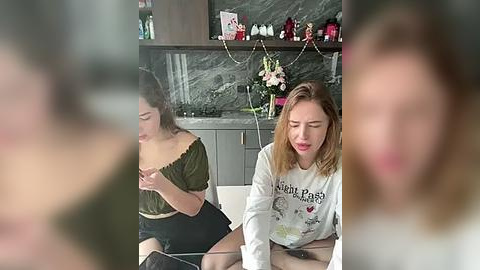 Media: Video of two women, one with long brown hair in a green off-shoulder top, and the other with blonde hair in a white graphic tee, sitting on a couch in a modern kitchen with dark countertops and decorative items.