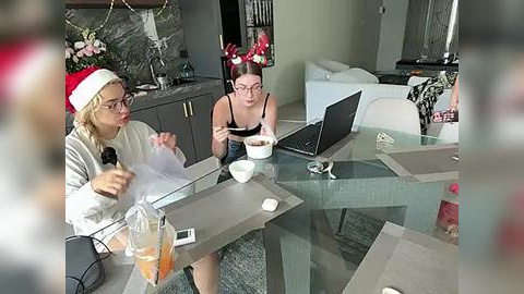 Media: Video of two women in a modern living room. One wears a Santa hat, glasses, and a sweater, while the other wears glasses and a black top. They sit at a glass table, working on laptops.