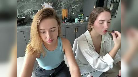 Media: Video of two young women, one applying lipstick, the other sitting, in a modern kitchen with grey marble countertops.