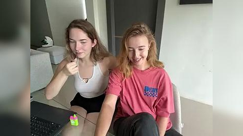 Media: Video of two young women, one in a white tank top, the other in a pink shirt, sitting on a black chair, both smiling. Background includes a mirror, white walls, and a desk with a green plant.