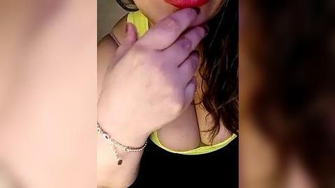 Media: Video of a woman with fair skin and long brown hair, wearing a lime green top, biting her finger, with a silver bracelet on her wrist.