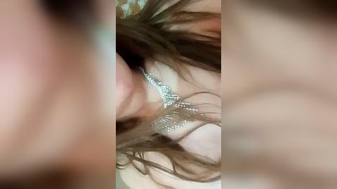 Media: A blurry video captures a close-up of a woman's face, focusing on her left ear adorned with a silver, intricately designed earring. Her long, wavy brown hair partially covers her face.