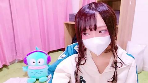 Media: Video of an Asian woman with long brown hair, wearing a white face mask, blue jacket, and pink headphones, sitting on a green carpet, next to a blue, cartoonish stuffed toy, in a pink-draped room.