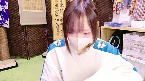 Media: Video of an Asian woman in a white surgical mask, wearing a blue and white jacket, sitting on a green rug in a cluttered, traditional Japanese room with tatami mats, paper scrolls, and various fabrics.