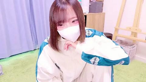 Media: Video of an Asian woman with shoulder-length straight brown hair, wearing a white and teal jacket with a shark motif, a white face mask, and a white hood, kneeling on a green carpet in a room with light-colored walls and a wooden ladder.