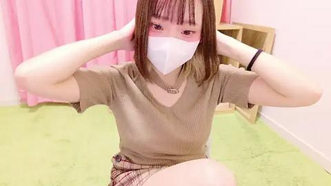 Media: Video of an East Asian woman with straight, shoulder-length brown hair, wearing a beige ribbed t-shirt and plaid shorts. She has a white face mask on and is adjusting her hair. Background features light green carpet and pink curtains.