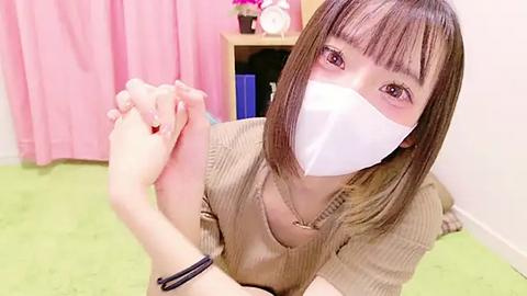 Media: Video of a young Asian woman with straight brown hair, wearing a white face mask and beige ribbed sweater, holding hands with another person, in a room with pink curtains and green carpet.