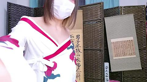 Media: Video of an Asian woman with shoulder-length brown hair, wearing a white kimono with red accents, a face mask, and standing in front of a Japanese folding screen with calligraphy and artwork.