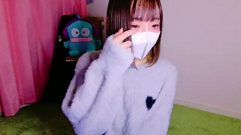 Media: Video of an East Asian woman with straight, shoulder-length brown hair, wearing a white fuzzy robe with a black heart symbol, and a white mask, kneeling on a green carpet in a room with pink curtains and a plush toy.