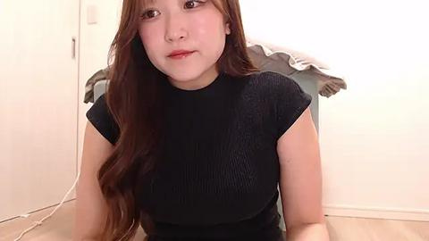 Media: A video of an East Asian woman with long, wavy brown hair, fair skin, and a petite physique, wearing a black ribbed top. She's sitting in a plain, softly lit room with a white wall and a piece of gray clothing hanging.