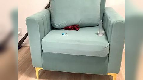 Media: Video of a teal armchair with a red cloth on its seat and a clear glass on its armrest, placed on a wooden floor in a minimalist room with white walls.