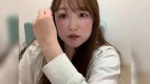 Media: Video of an East Asian woman with long brown hair, fair skin, and a neutral expression, resting her head on her left hand. She wears a white jacket, seated against a blurred background of a chair and light-colored wall.