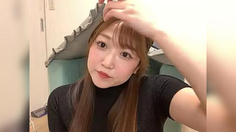 Media: Video of a young Asian woman with straight, light brown hair, wearing a black turtleneck, sitting in a teal chair, adjusting her hair. Background shows a white wall and a ruffled pillow.