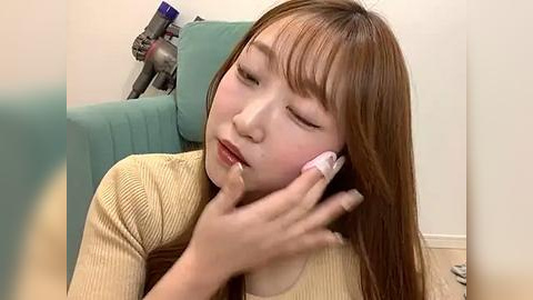 Media: Video of a young Asian woman with long, straight brown hair, wearing a beige ribbed sweater, talking on a white phone. She sits on a green couch against a light beige wall.