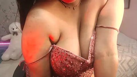 Media: Video of a woman with fair skin and dark hair, wearing a low-cut, sequined, pink dress. Her cleavage is prominent, and she is in a bedroom with a white teddy bear and a geometric-patterned bedspread.