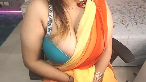 Media: Video of a woman in a yellow saree with a blue bra visible, wearing a gold bracelet, seated in a modern room with a floral-patterned bed and a blue pool in the background.