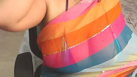 Media: Video of a woman's bare chest, partially covered by a vibrant, multicolored, striped, and sequined scarf. She has light skin and is wearing a black bra.