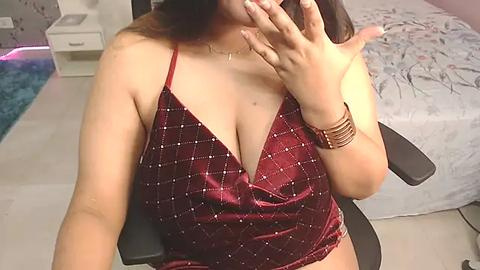 Media: Video of a plus-sized woman with medium skin tone, wearing a plunging maroon velvet dress, sitting in a black office chair. She covers her face with one hand, revealing manicured nails. Background includes a bed with floral bedding and a nightstand.