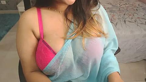 Media: Video of a light-skinned woman with long, dark hair, wearing a pink bra and a blue, translucent robe, sitting on a bed with floral bedding.