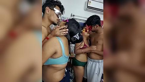 Media: Video of a shirtless man in a blue bra, kissing a woman in a red bikini, with a third person in the background, in a messy room.