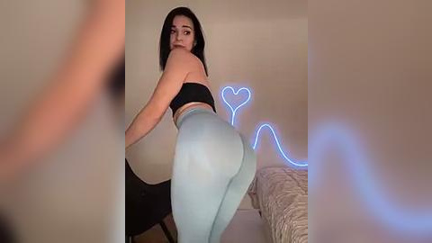 Media: A video of a curvaceous, light-skinned woman with long dark hair, wearing a black bra and light blue leggings, bending over, highlighting her large buttocks. She stands in a softly lit, beige room with neon heart decorations.