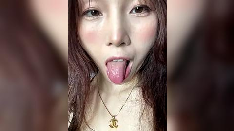 Media: A close-up video of an East Asian woman with fair skin and long, straight, brown hair. She is sticking out her tongue, wearing a gold necklace with a small pendant. The background is blurred.
