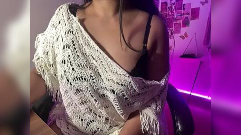 Media: Video of a woman with medium brown skin, wearing a white crocheted shawl over a black bra, sitting in a dimly lit room with purple ambient lighting, a wall collage, and a black chair.