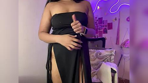 Media: Video of a woman with medium skin tone, wearing a black strapless dress with high slit, holding a phone. Background features a bed with patterned pillows and neon lights.