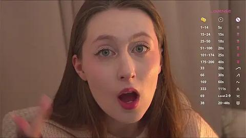 Media: Video of a young Caucasian woman with light brown hair, green eyes, and fair skin, wearing a beige sweater, looking surprised and open-mouthed. Text overlay with \"Catherine\" and \"1:14\" at the top.