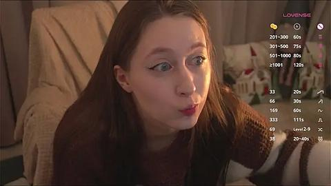 Media: Video of a young Caucasian woman with fair skin and long brown hair, wearing a brown sweater, blowing a kiss. Background includes a beige curtain and a toy house.