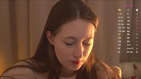 Media: Video of a young Asian woman with long brown hair, wearing a beige sweater, gazing downward in a softly lit, indoor setting.