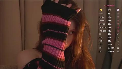 Media: A video showing a woman with long, wavy brown hair, partially obscured by a black-and-pink striped knit hat, in a dimly lit room.