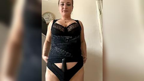 Media: A video of a plus-size woman with light skin, dark hair, and tattoos, wearing black lingerie with lace and a black dildo, standing in a dimly lit room with a clock and wall decor.