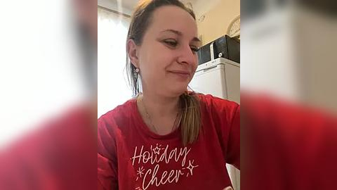 Media: A video of a smiling woman with light skin, brown hair tied back, wearing a red \"Holiday Cheer\" shirt. Background includes a white fridge and microwave, blurred.